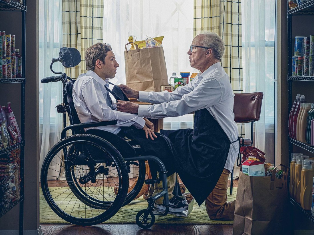 An older man adjusting the tie of a young man in a wheelchair, surrounded by grocery shelves, from a live-action film